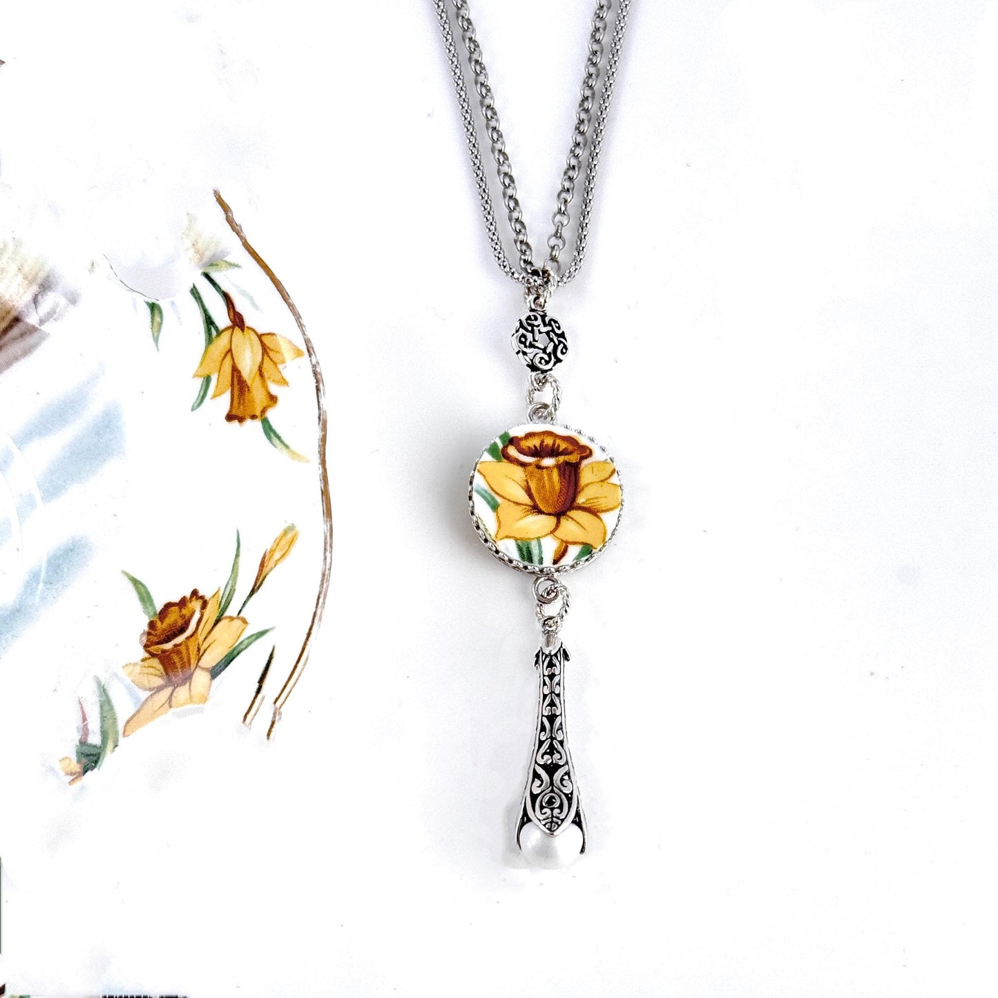 May include: A silver chain necklace with a round porcelain pendant featuring a yellow daffodil. The pendant is attached to a silver charm with a pearl hanging from it.
