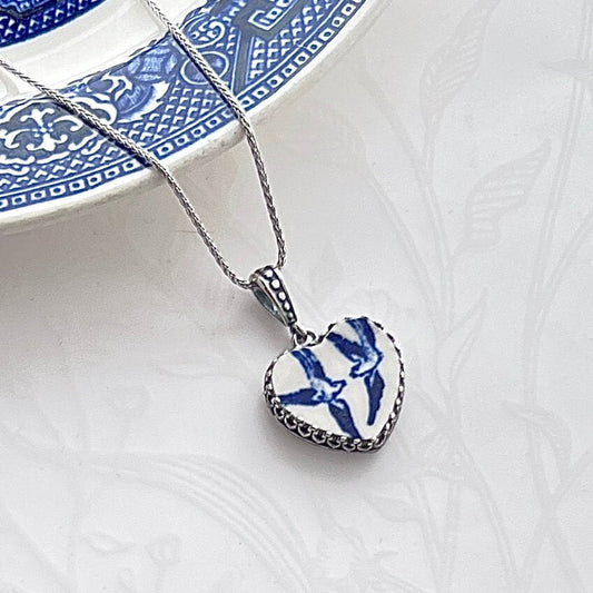 May include: A silver chain necklace with a heart-shaped pendant. The pendant is made of white porcelain with a blue and white design of two love birds in flight.