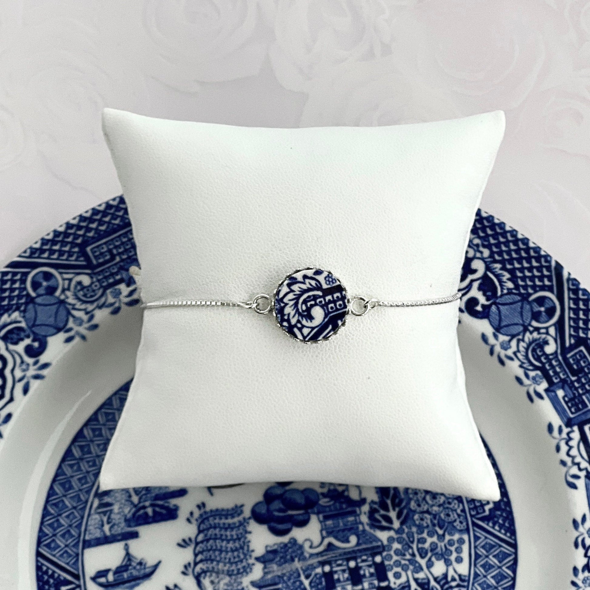 May include: A delicate bracelet featuring a circular blue and white ceramic charm. The charm showcases a detailed floral pattern reminiscent of vintage blue and white china. The bracelet has a thin, silver chain. The bracelet is displayed on a white pillow resting on a blue and white china plate, enhancing the overall theme of classic elegance and timeless style.