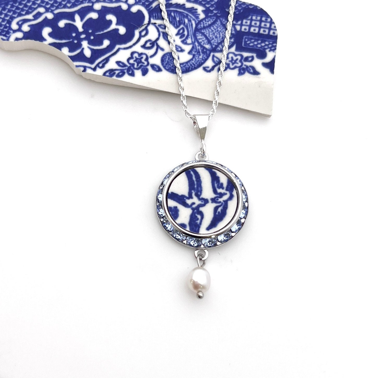 Necklace with a handmade round pendant that features love birds on a vintage blue willow china pattern. Sparkly crystals surround the china in a silver-tone frame. Pendant is 1 inch round with a sterling silver chain, loop style bail and pearl drop.