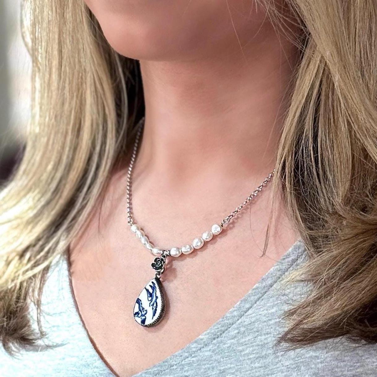May include: A silver chain necklace with a white pearl strand and a teardrop-shaped pendant. The pendant is blue and white with a floral design.