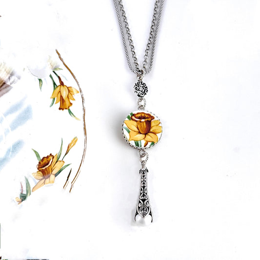 May include: A silver chain necklace with a round porcelain pendant featuring a yellow daffodil. The pendant is attached to a silver charm with a pearl hanging from it.