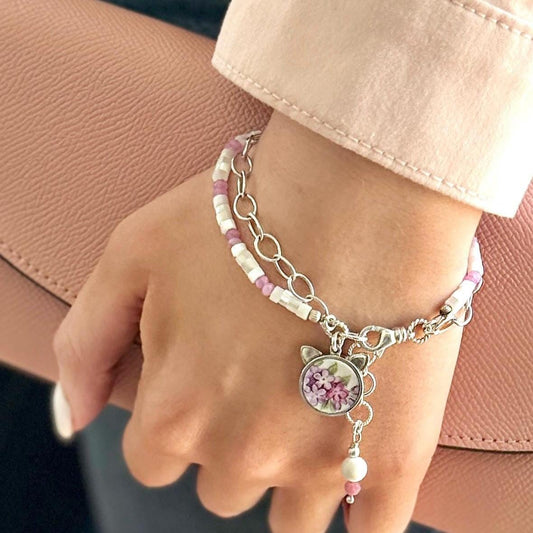 May include: A silver chain bracelet with a white and purple beaded bracelet layered on top. The bracelet has a charm with a floral design and a pearl dangle.