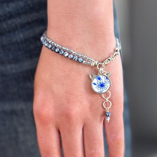May include: A silver bracelet with a blue and white floral charm. The charm has a cat face design with small silver ears. The bracelet is made of small silver beads and has a silver chain with a silver clasp.