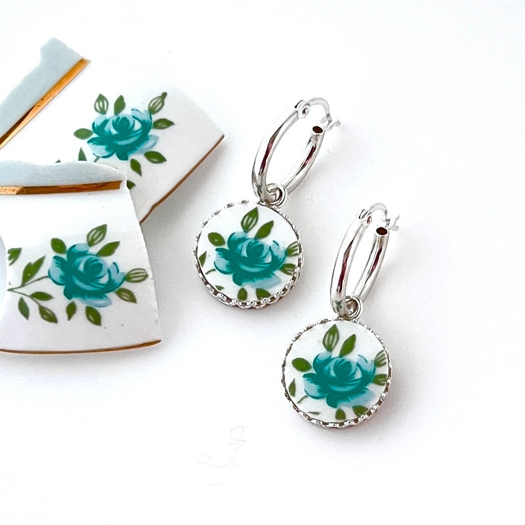 May include: A pair of silver hoop earrings with round, white pendants. Each pendant features a teal rose design with green leaves and a scalloped silver edge. The earrings are displayed on a white background, with pieces of broken white ceramic with teal rose designs visible.