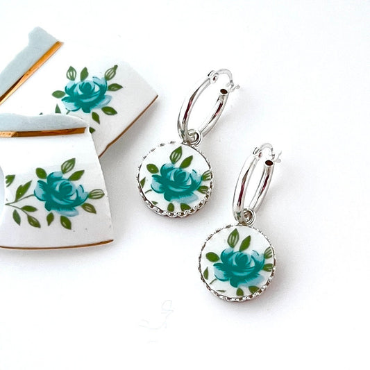 May include: A pair of silver hoop earrings with round, white pendants. Each pendant features a teal rose design with green leaves and a scalloped silver edge. The earrings are displayed on a white background, with pieces of broken white ceramic with teal rose designs visible.