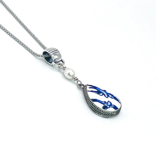 May include: A silver chain necklace with a teardrop-shaped pendant. The pendant is white with a blue floral design and a silver border. A single white pearl is attached to the chain above the pendant.