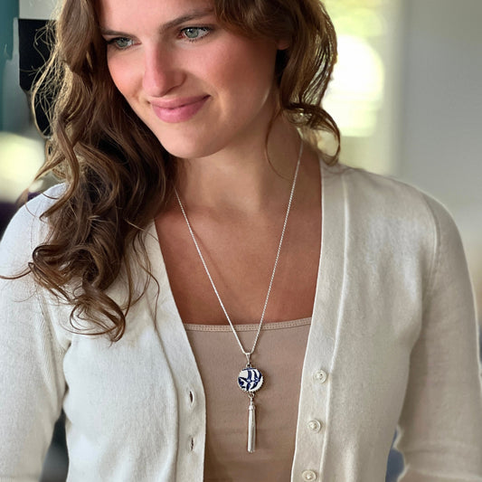 May include: A silver necklace with a blue and white patterned circular pendant and a silver tassel. The necklace is on a white cardigan.