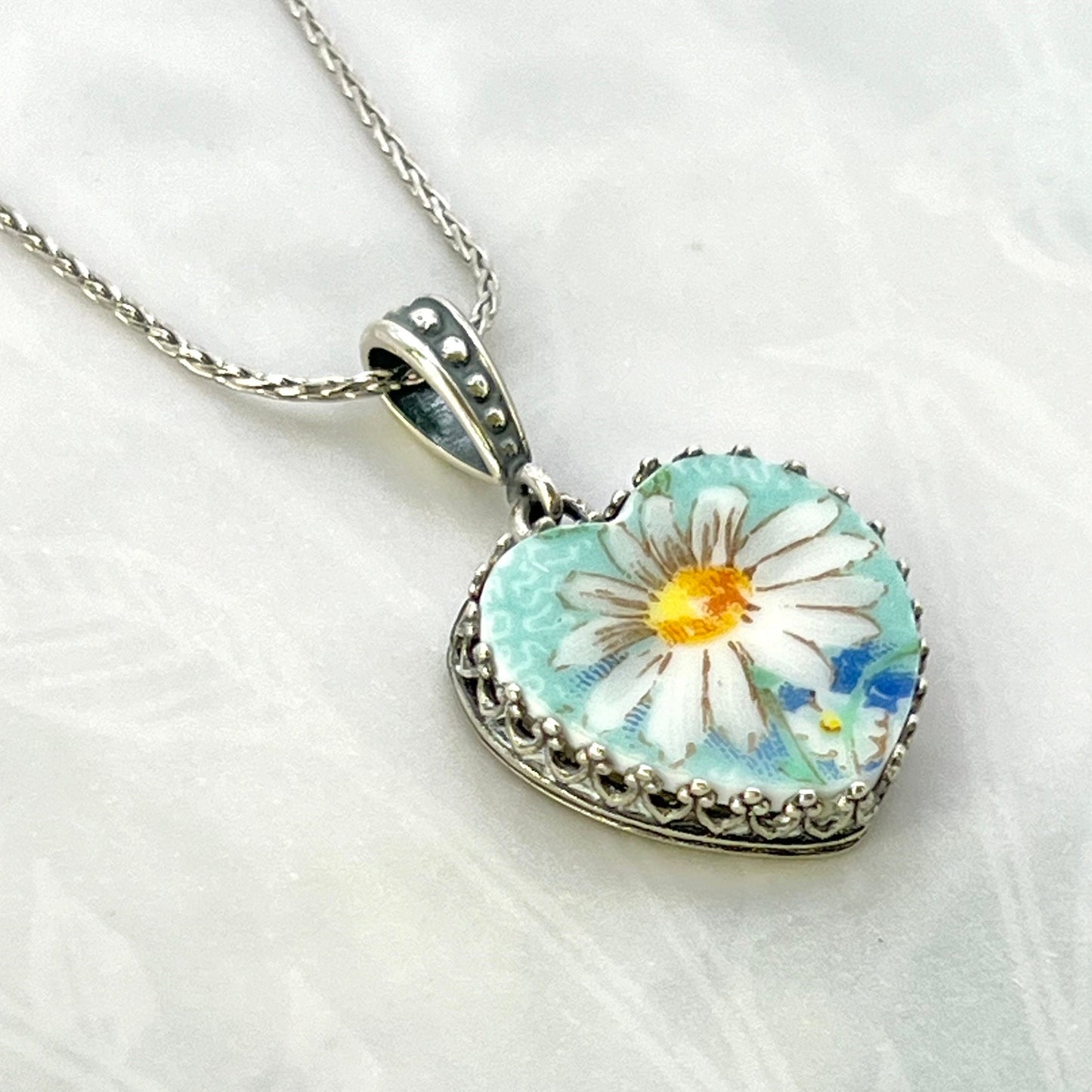 May include: A silver chain necklace with a heart-shaped pendant. The pendant features a white daisy with a yellow center on a light blue background. The pendant is surrounded by a silver filigree border.