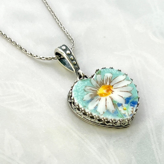 May include: A silver chain necklace with a heart-shaped pendant. The pendant features a white daisy with a yellow center on a light blue background. The pendant is surrounded by a silver filigree border.
