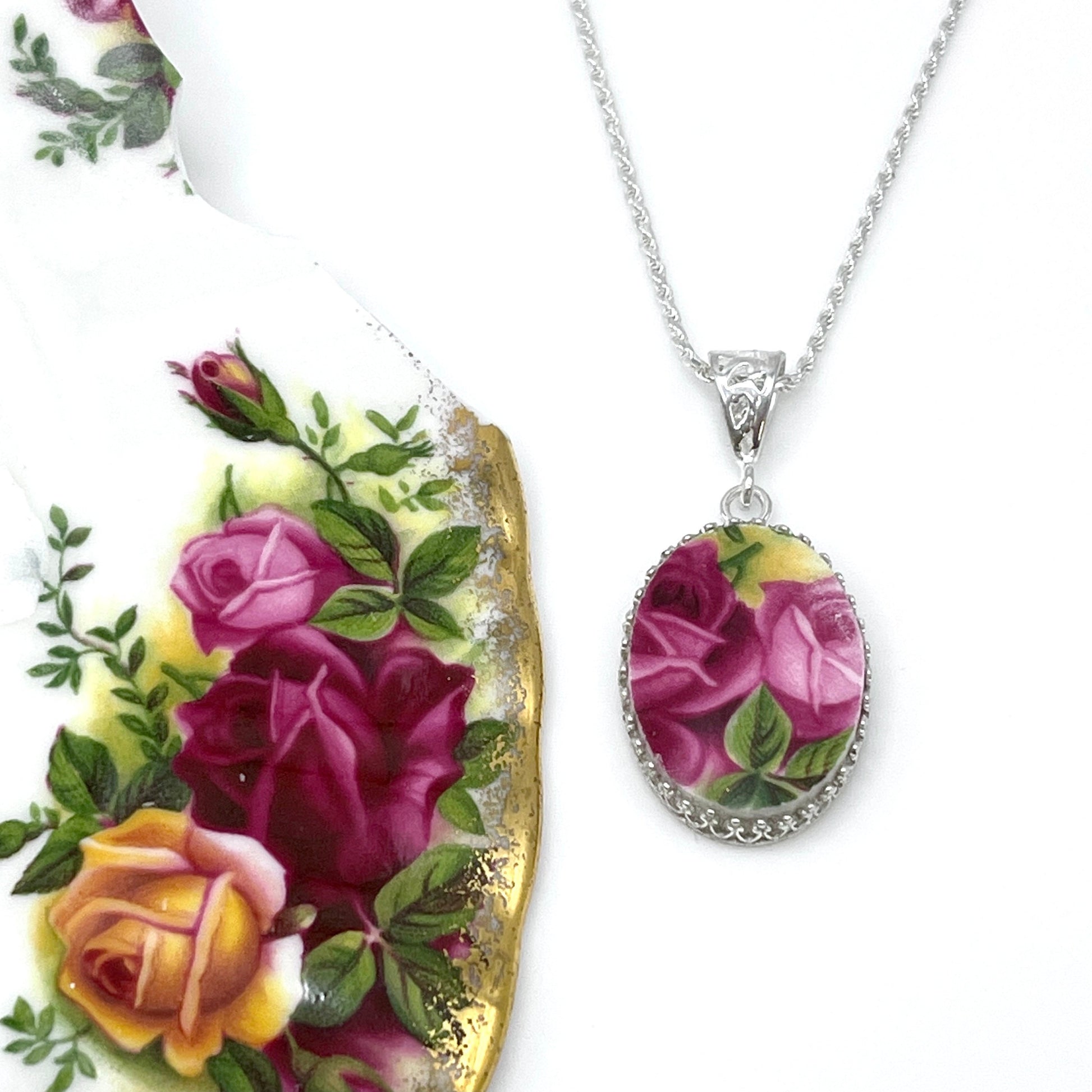 May include: A silver chain necklace with an oval pendant featuring a floral design. The pendant is made of porcelain and features a pink and white rose pattern on a cream background. The pendant is surrounded by a silver border with a decorative edge.