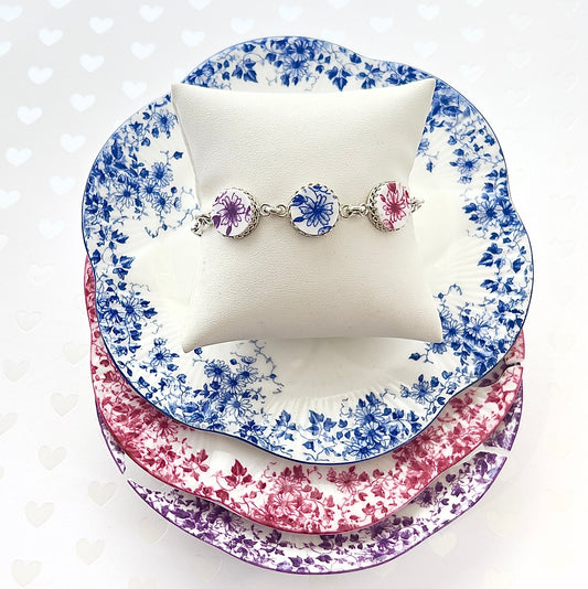 May include: A silver bracelet with three circular charms, each featuring a floral design. The flowers are in shades of purple and blue against a white background. The bracelet is displayed on a white cushion, with three decorative plates in blue, red, and purple floral patterns stacked beneath.
