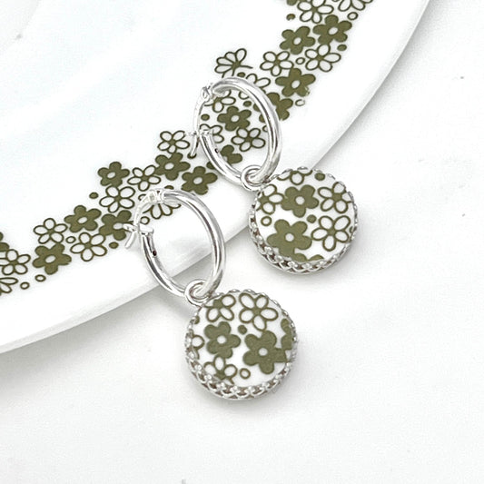 May include: A pair of silver hoop earrings with a white and green floral design. The earrings are hanging from a white surface.
