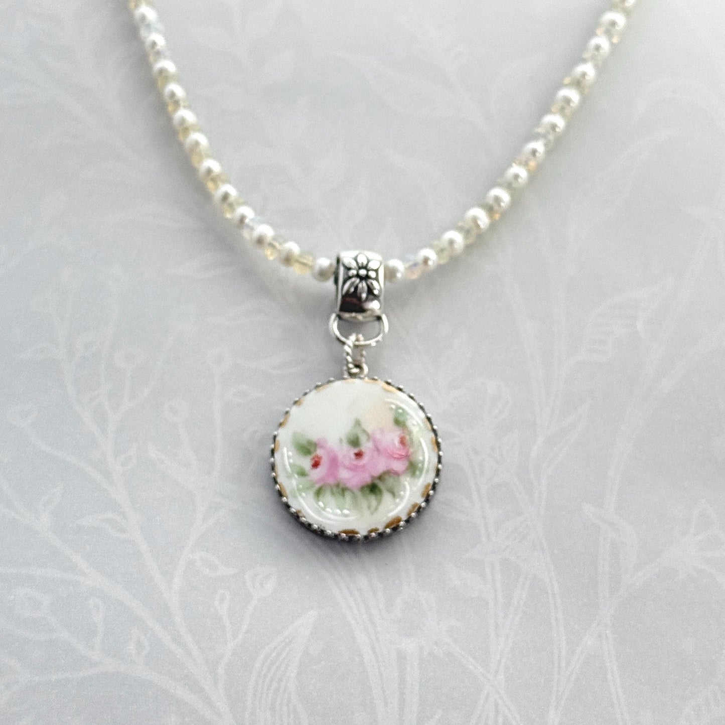 May include: A delicate necklace featuring a round pendant with a floral design. The pendant showcases pink roses with green leaves on a white background, framed by a silver-colored beaded edge. The necklace has a beaded chain with small, light-colored beads and a decorative clasp.