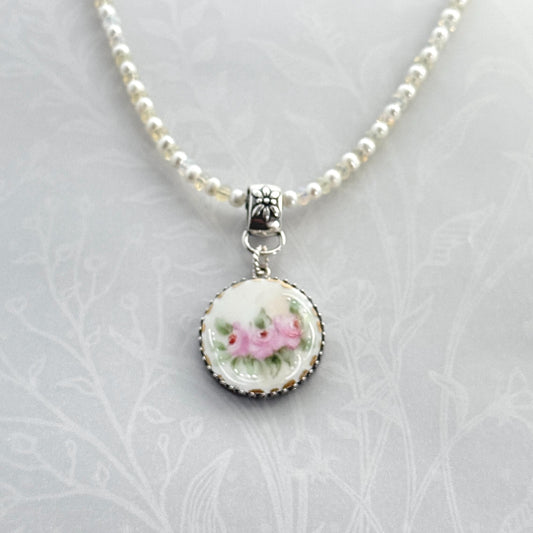 May include: A delicate necklace featuring a round pendant with a floral design. The pendant showcases pink roses with green leaves on a white background, framed by a silver-colored beaded edge. The necklace has a beaded chain with small, light-colored beads and a decorative clasp.