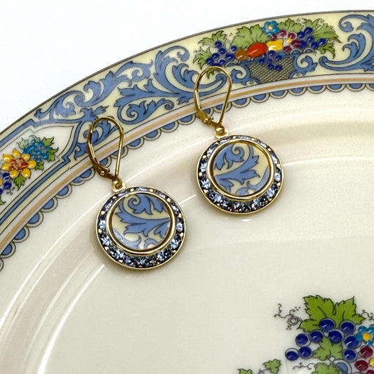 May include: A pair of gold tone earrings with a blue and white floral design. The earrings are round and have a circle of clear rhinestones around the edge.