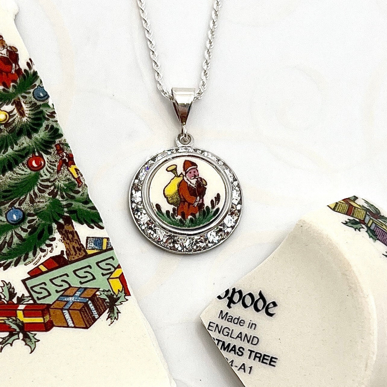 May include: A silver necklace with a round pendant featuring a white porcelain image of a man carrying a sack on his back. The pendant is surrounded by a circle of clear rhinestones. The pendant is attached to a silver chain. The necklace is made by Spode and says &#39;Made in England Christmas Tree 24-A1&#39;.