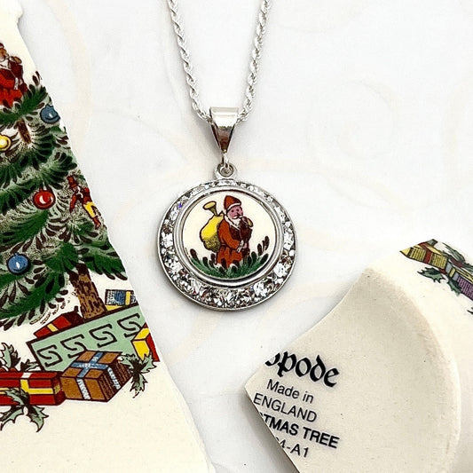 May include: A silver necklace with a round pendant featuring a white porcelain image of a man carrying a sack on his back. The pendant is surrounded by a circle of clear rhinestones. The pendant is attached to a silver chain. The necklace is made by Spode and says &#39;Made in England Christmas Tree 24-A1&#39;.