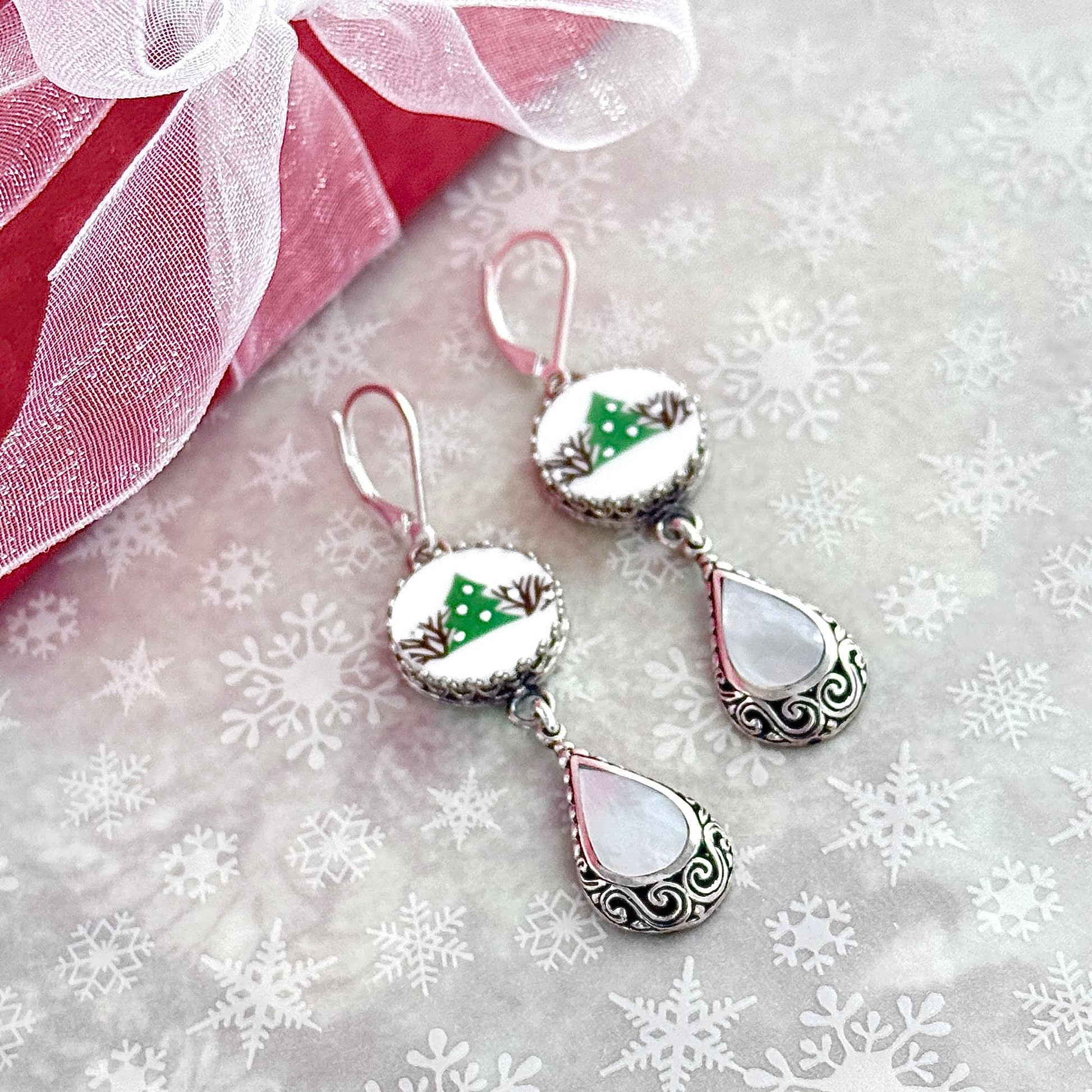 May include: Silver Christmas earrings with white and green accents. Each earring features a round cabochon with a small green Christmas tree design on a white background, followed by a pearlescent teardrop charm with silver filigree detailing. The earrings have a dangling design and are suitable for holiday wear.