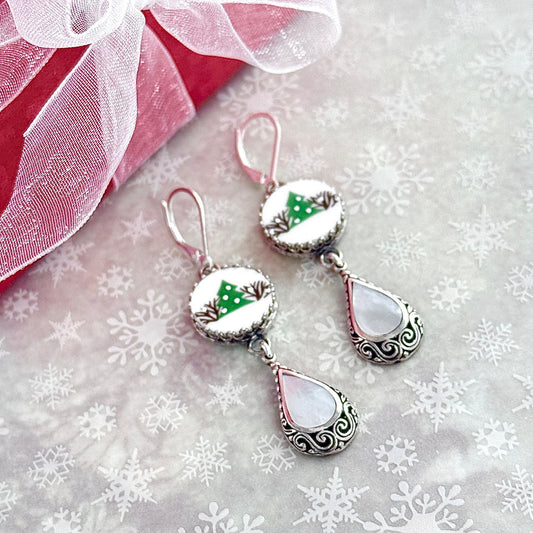 May include: Silver Christmas earrings with white and green accents. Each earring features a round cabochon with a small green Christmas tree design on a white background, followed by a pearlescent teardrop charm with silver filigree detailing. The earrings have a dangling design and are suitable for holiday wear.