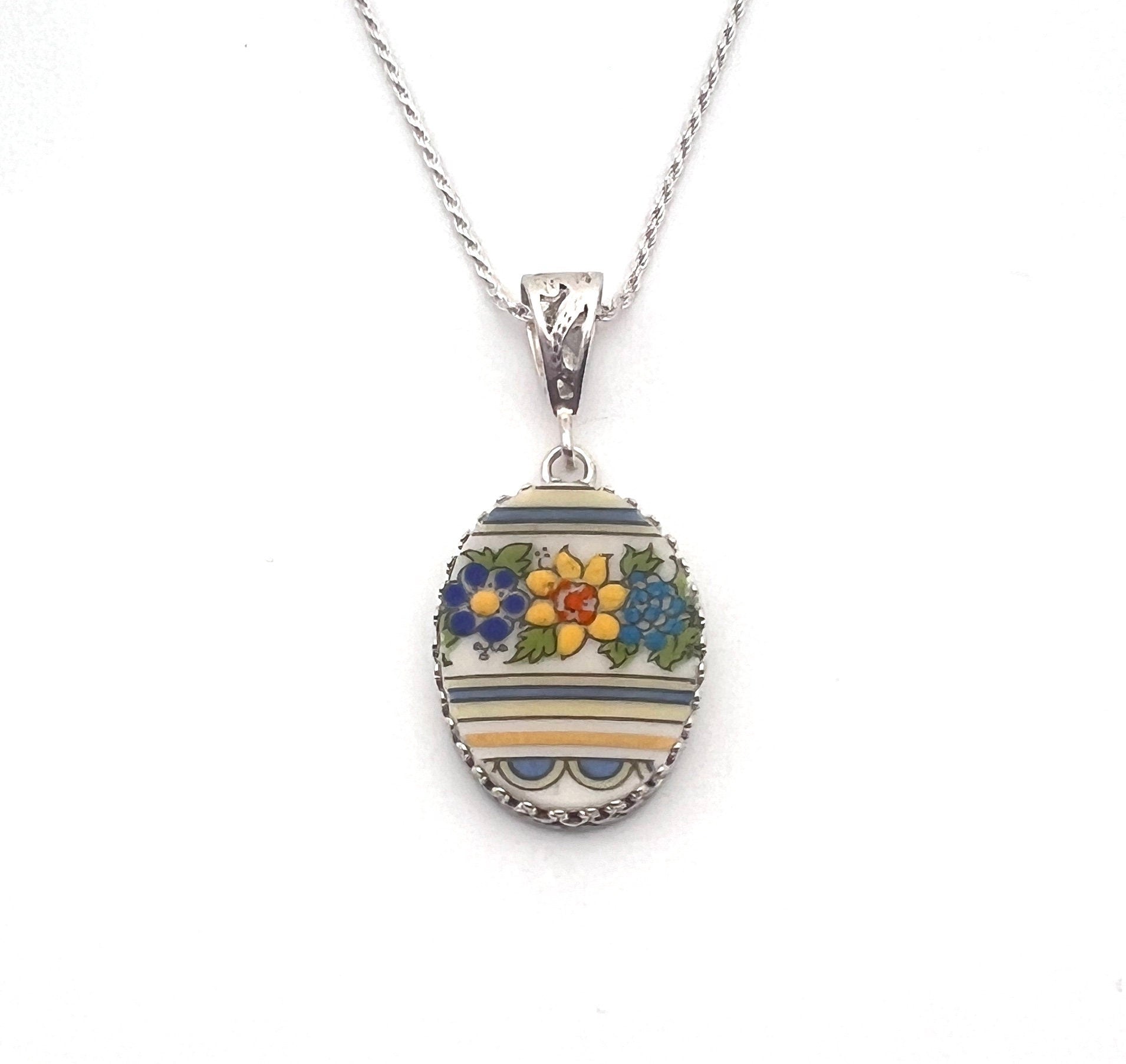 May include: A silver chain necklace with an oval pendant. The pendant is white with blue and yellow stripes and a floral design in blue, yellow, orange, and green. The pendant is surrounded by a silver border.