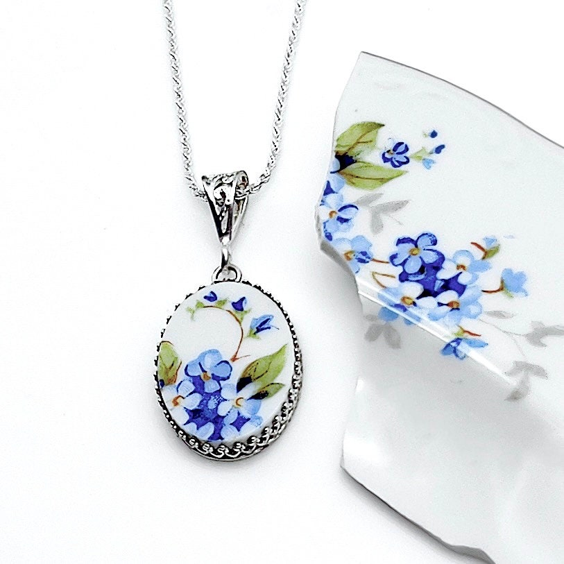 May include: A silver chain necklace with an oval pendant featuring a blue and green floral design on a white background. The pendant is surrounded by a silver beaded border.