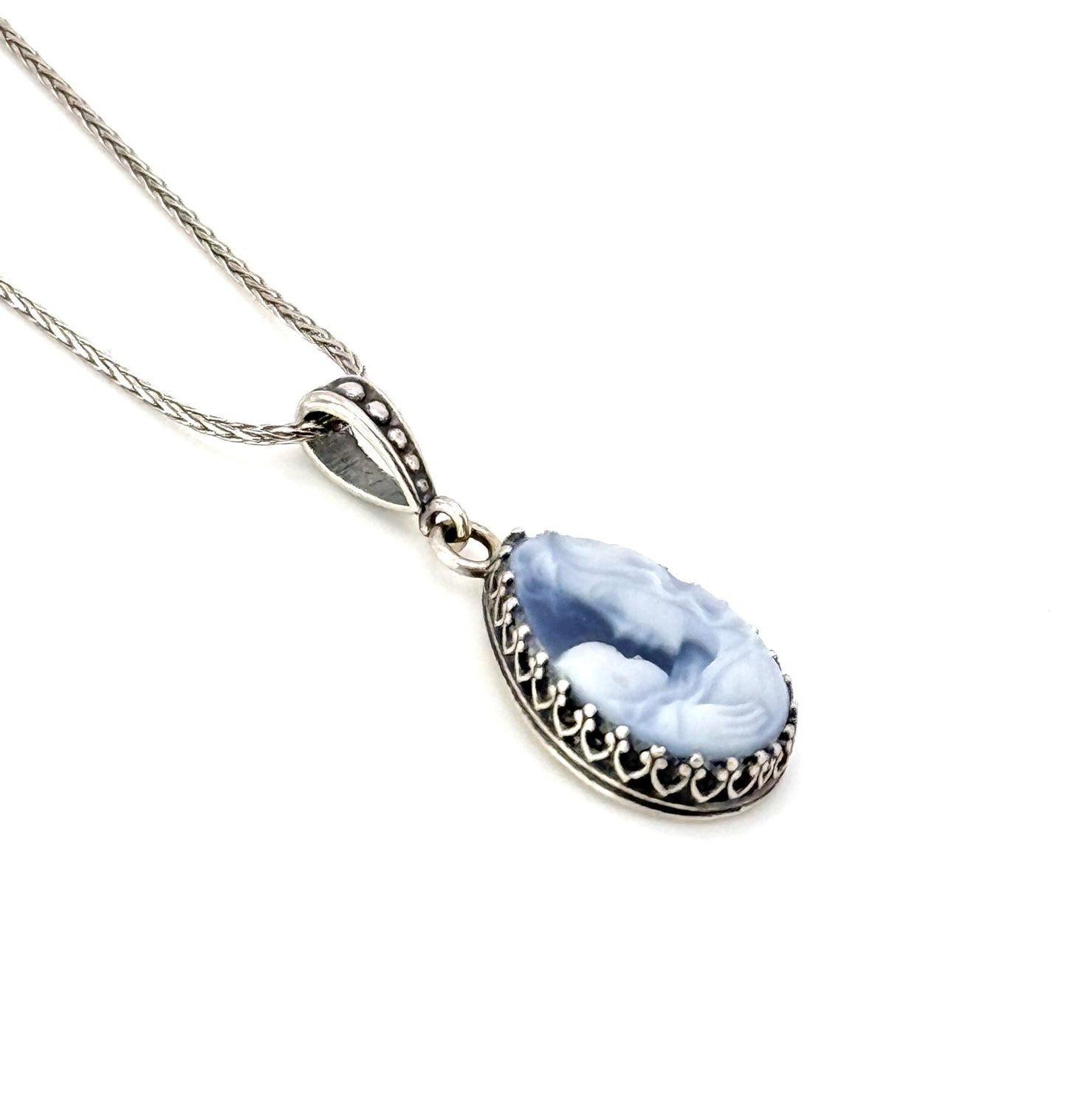 May include: A silver chain necklace with a teardrop-shaped pendant. The pendant is made of a light blue stone with a carved design and a silver border.