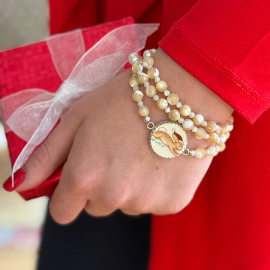 May include: Layered beige and off-white pearl beaded bracelets with a charming rabbit charm pendant. The rabbit charm is set against a cream-colored background and features a red Santa hat. The bracelets are elegantly designed and perfect for adding a touch of festive flair to any outfit.