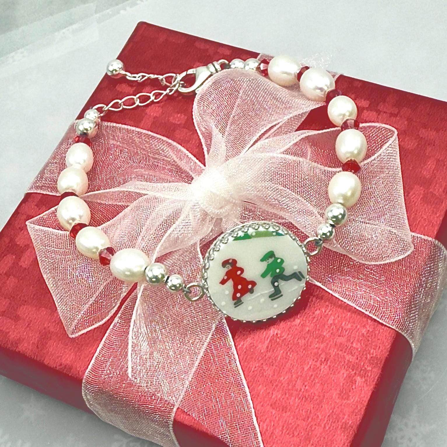 May include: A pearl and crystal bracelet features a silver clasp and a circular pendant with a charming illustration of children ice-skating. The pendant is white with green and red accents. The bracelet is presented on a red gift box adorned with a white ribbon, creating a festive and elegant presentation. The overall aesthetic is one of winter holidays and festive cheer.