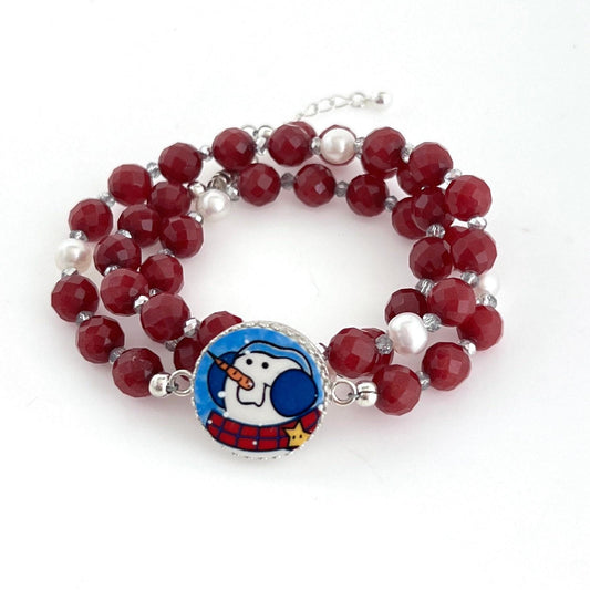 May include: A triple-wrap bracelet features faceted burgundy red gemstone beads interspersed with small silver beads and white pearls. A circular silver charm with a whimsical snowman illustration is attached to the bracelet. The snowman is depicted in blue and white, wearing a red and black plaid scarf, and has a carrot nose. The bracelet is a festive accessory, perfect for winter holidays.