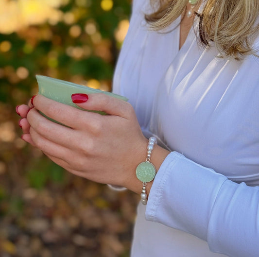 May include: A delicate bracelet features a light green jade disc pendant encircled by small white pearls. The bracelet has a silver-toned metal chain and clasp.  The jade disc has an etched floral pattern. The bracelet is shown on a wrist, alongside a pale green ceramic bowl.