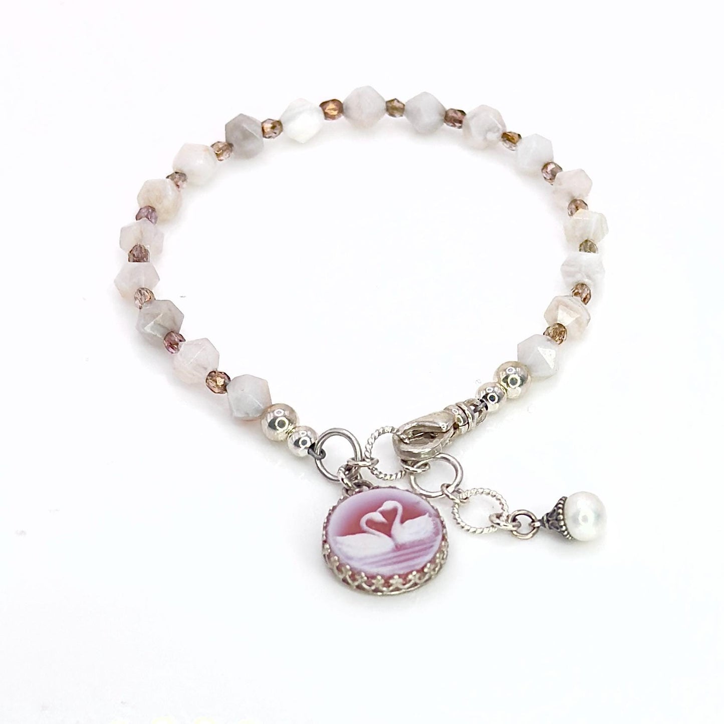 May include: A delicate bracelet featuring a series of faceted, light-colored beads. A round, pink cameo pendant with two swans is attached, along with a small pearl charm. The bracelet is secured with a silver clasp.