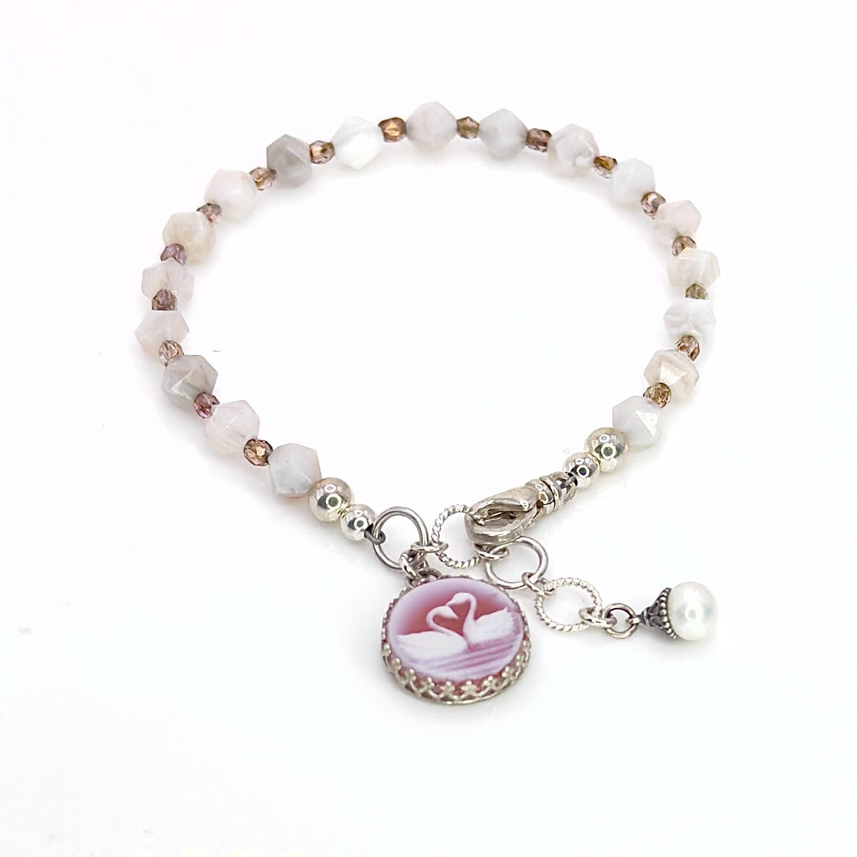 May include: A delicate bracelet featuring a series of faceted, light-colored beads. A round, pink cameo pendant with two swans is attached, along with a small pearl charm. The bracelet is secured with a silver clasp.