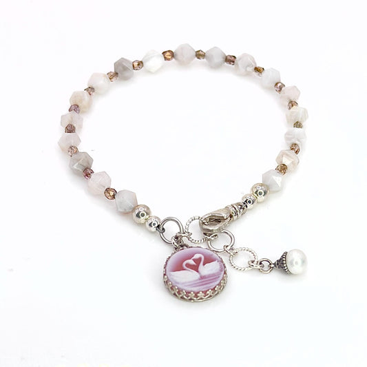 May include: A delicate bracelet featuring a series of faceted, light-colored beads. A round, pink cameo pendant with two swans is attached, along with a small pearl charm. The bracelet is secured with a silver clasp.