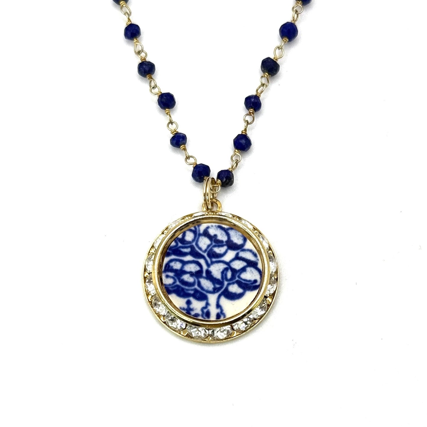 May include: A gold-colored necklace with a round pendant. The pendant has a white background with a blue floral design. The pendant is surrounded by small, clear gemstones. The necklace chain is made of small, dark blue beads.