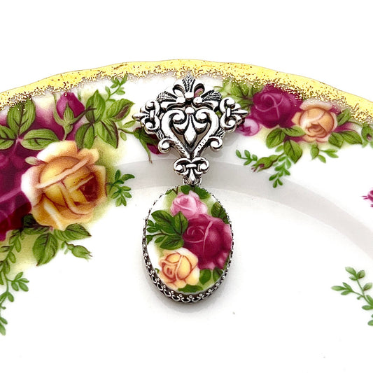 May include: Oval-shaped pendant featuring a cameo of pink, yellow, and burgundy roses with green leaves. The cameo is framed by a silver-toned metal border with a detailed filigree bail. The pendant is displayed on a plate with a floral pattern.