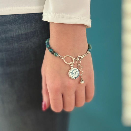 May include: A bracelet featuring a strand of teal and brown beads, connected to a silver chain. A circular charm with a blue and white design hangs from the chain, along with a small dangling pearl. The bracelet is fastened with a silver clasp.