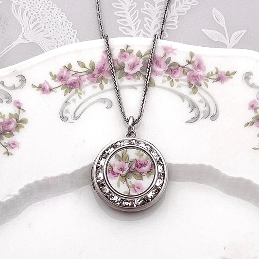 May include: A silver locket necklace with a white porcelain cameo featuring pink roses. The locket is surrounded by clear rhinestones.