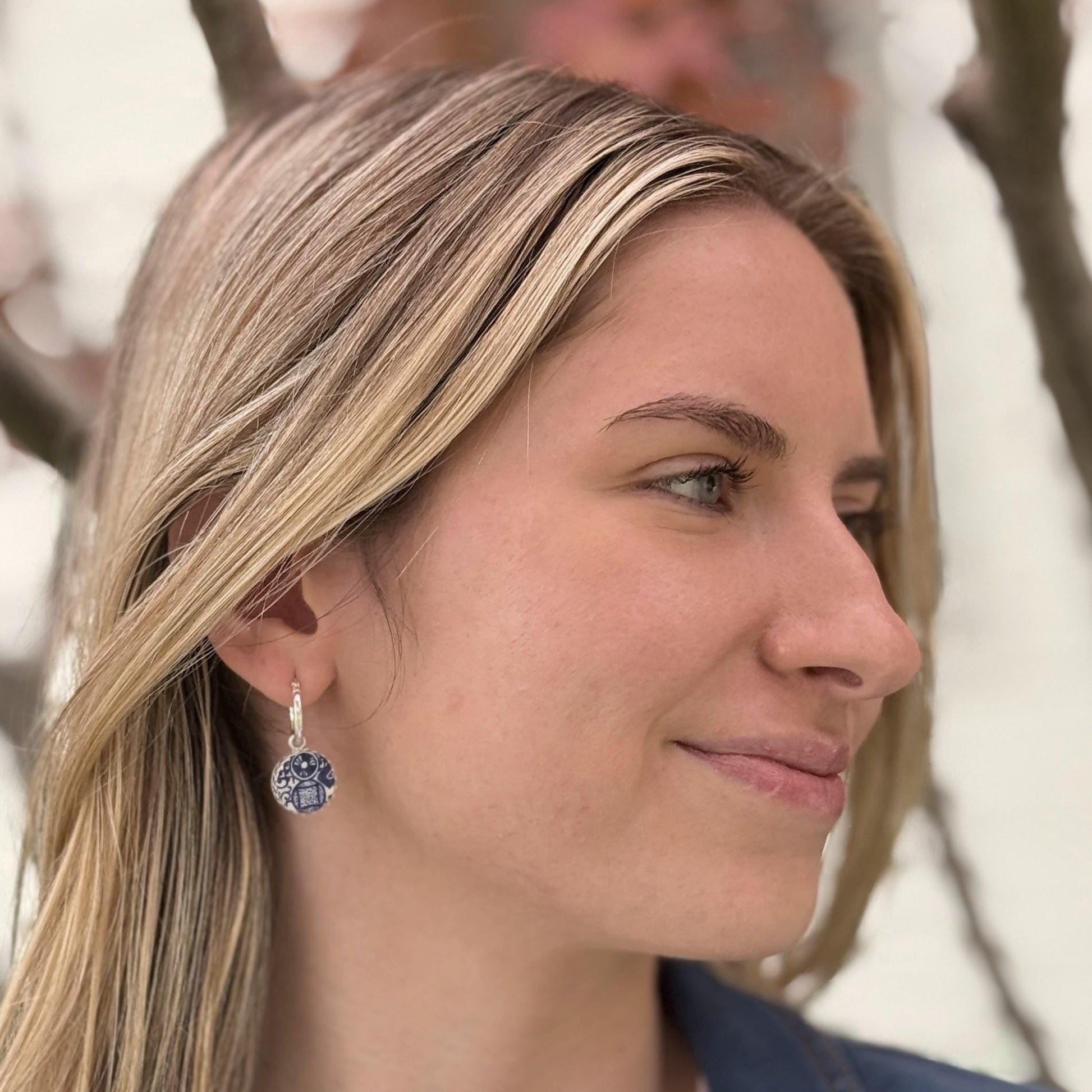 May include: Silver hoop earrings with a round, blue and white pendant. The pendant features a geometric design with a central square and surrounding details. The earrings are worn by a person with blonde hair.