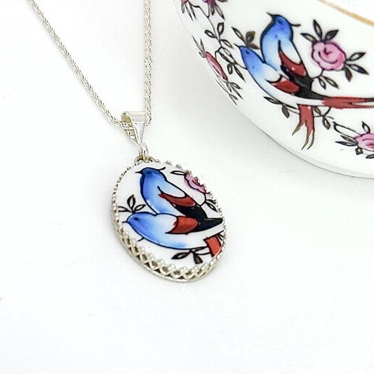May include: Silver-toned necklace with an oval pendant featuring a hand-painted design of two blue birds with red and black accents, surrounded by pink flowers and green leaves. The pendant has a decorative, openwork silver border.