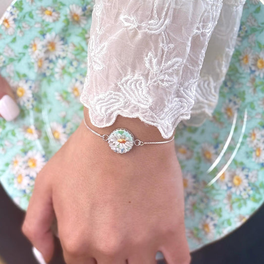 May include: A silver chain bracelet with a white daisy charm made of mosaic tiles. The charm is surrounded by a silver frame.