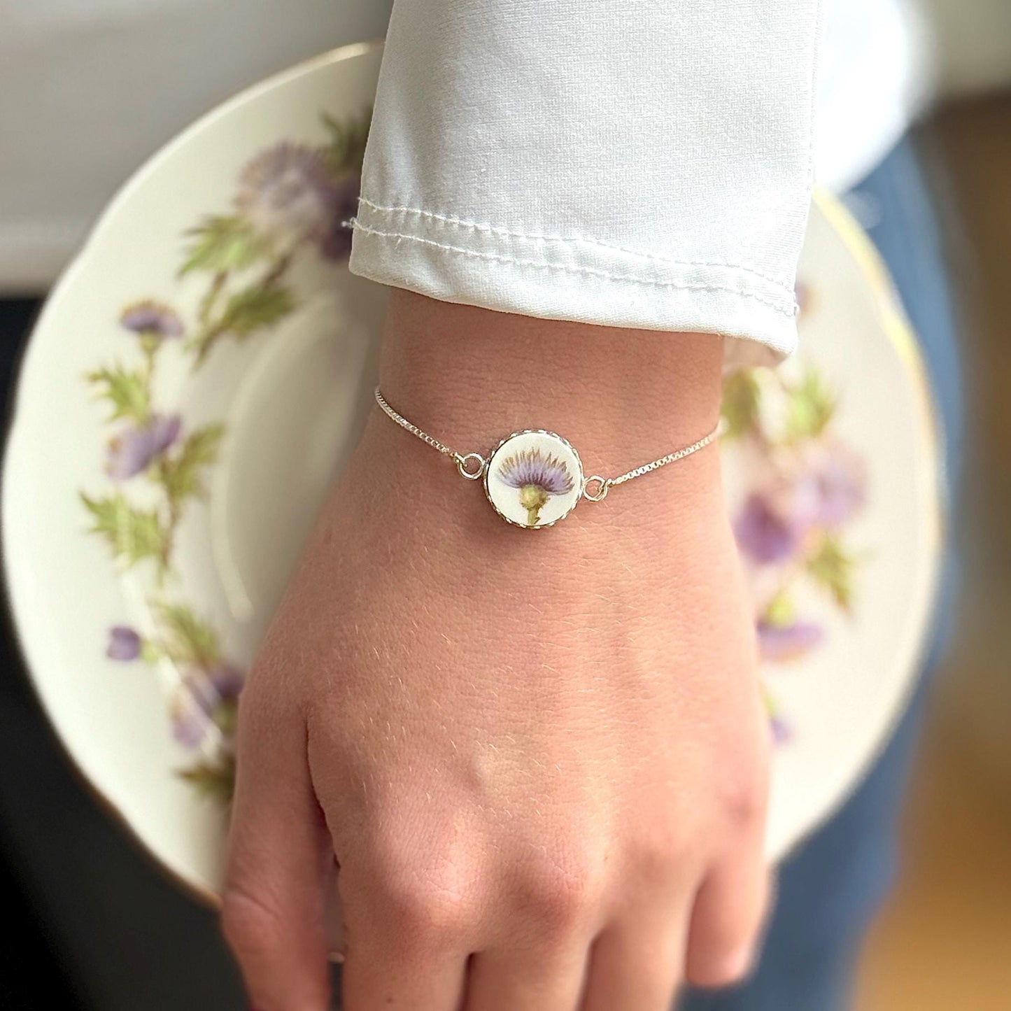 May include: Silver bracelet featuring a round, bezel-set pendant with a pressed purple flower. The delicate chain bracelet is worn on a wrist, with a floral-patterned plate in the background. The bracelet's pendant has a white border.