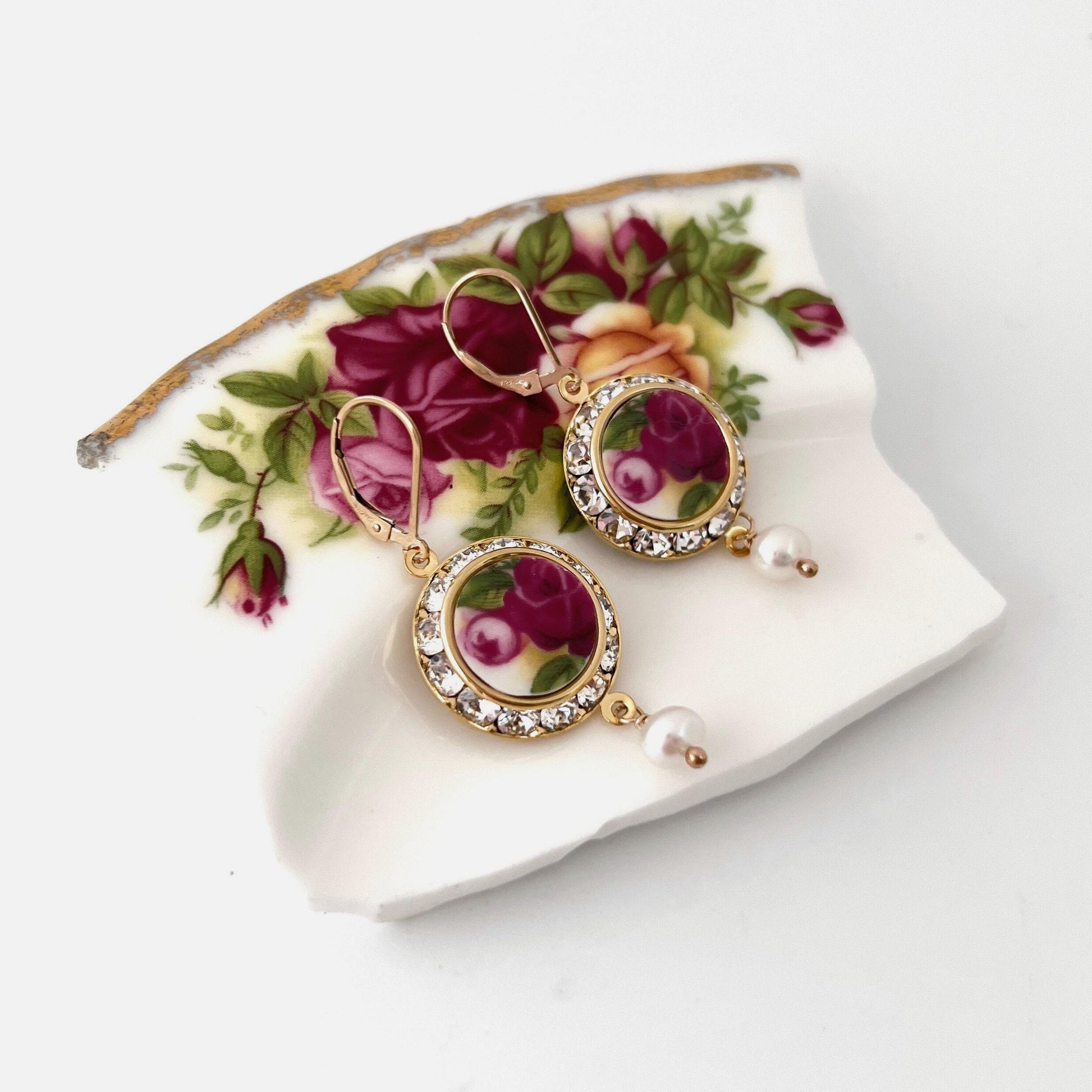 May include: A pair of gold earrings with a round, white porcelain base with a pink floral design. The porcelain base is surrounded by a ring of clear rhinestones. Each earring has a dangling pearl drop.