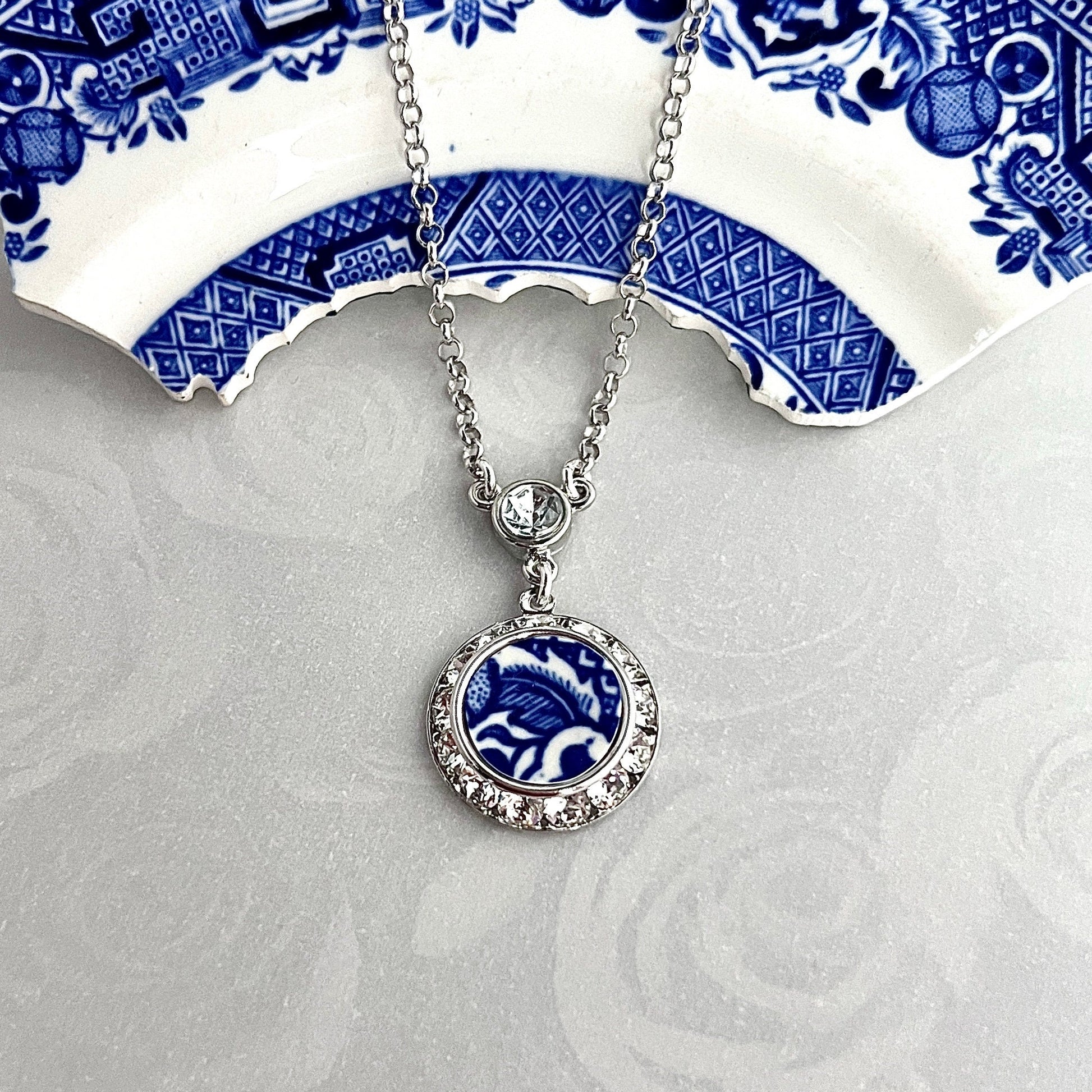 Necklace with a handmade round pendant that features the border pattern from a vintage blue willow china plate. Sparkly crystals surround the china in a silver-tone frame. Pendant measures ¾ inch round with a sterling silver chain and a crystal bail.