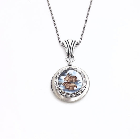May include: Silver-tone locket necklace with a round pendant featuring a painted image of two brown rabbits. The pendant is encircled with clear crystals and hangs from a delicate chain. The locket is a charming piece of jewelry.