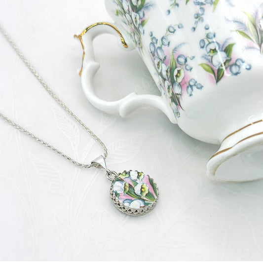 May include: A silver necklace with a round pendant featuring a floral design in pink, white, and green. The pendant is set in a decorative silver frame. A white teacup with a floral pattern is in the background.