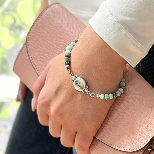 May include: A beaded bracelet with green and white stones and a decorative oval centerpiece featuring a floral design. The bracelet is silver-toned and is worn on a wrist, with a pink wallet in the background.