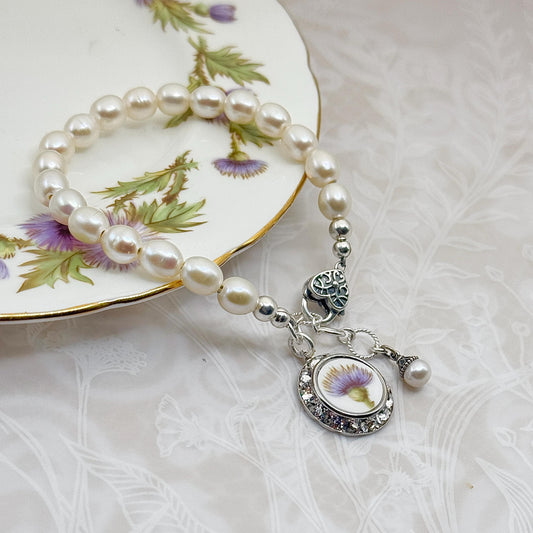 May include: A pearl bracelet with a silver charm featuring a purple thistle flower design. The bracelet is on a white plate with a gold rim and floral pattern. The charm is surrounded by small clear crystals.