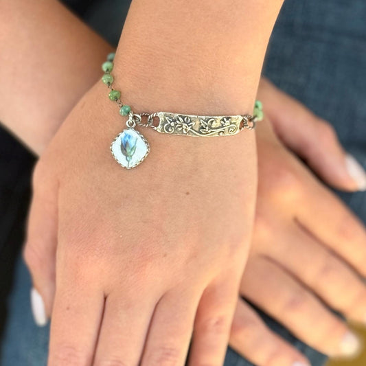 May include: A silver bracelet with a floral design and a small, square pendant featuring a blue flower. The bracelet has green beaded accents and is worn on a wrist.
