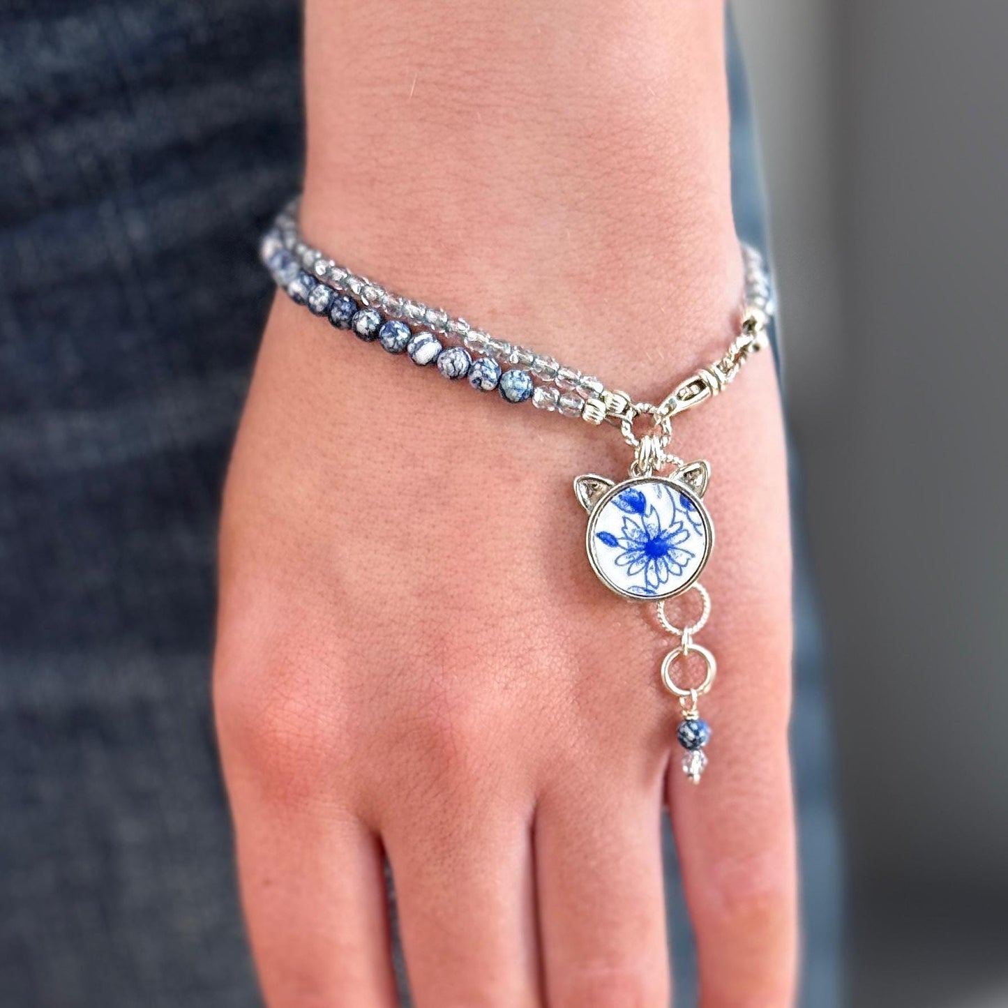 May include: A silver bracelet with a blue and white floral charm. The charm has a cat face design with small silver ears. The bracelet is made of small silver beads and has a silver chain with a silver clasp.